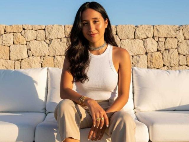AnnieBlakke with long dark hair, white sleeveless top, beige pants, gold jewelry, sitting on white couch, smiling, stone wall background, clear blue sky, warm sunlight.