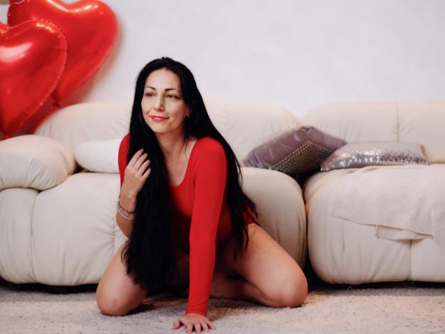 LiluGrants, long black hair, red long-sleeve top, kneeling on carpet, smiling, red heart balloons in background, white leather sofa, purple and silver pillows, red lipstick, bracelet.