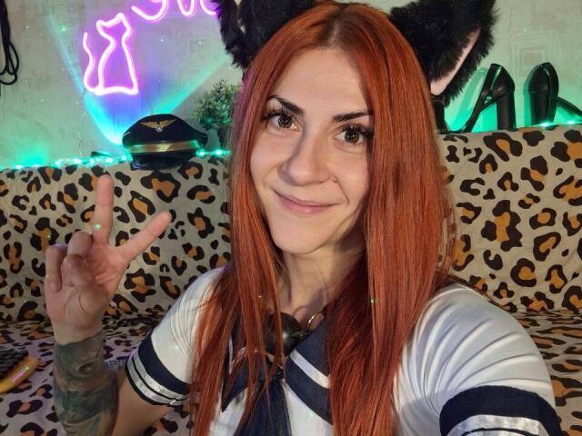 RedHurricane, a white woman with red hair and cat ears, smiling, wearing a navy and white sailor outfit, making a rock-paper-scissors gesture, visible tattoo on right arm.