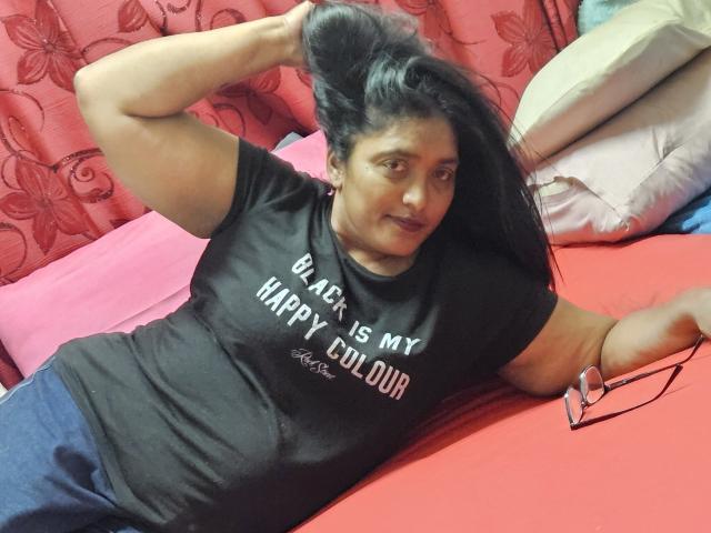 IndiaBlackPanther, dark-skinned woman with long black hair, lying on red bed, black t-shirt, holding hair, glasses on bed, blue pants, slightly smiling, hand on head.