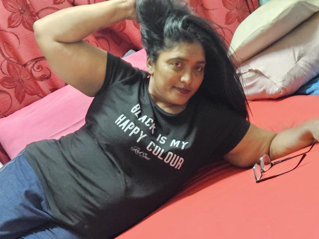 IndiaBlackPanther, a plus-sized woman with long black hair, lies on a red bed, wearing a black shirt and blue pants, with glasses on the bed and a pillow behind.