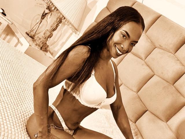 JadaMillss with dark skin, long straight hair, smiling, wearing white lace bra and panties, sitting on bed with tufted headboard, hands on bed, showing her tits and pussy.
