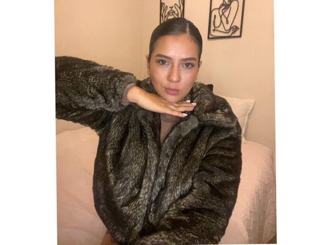 Velka in a furry green coat, hand on neck, looking at camera, brown skin, black hair in a ponytail, small ass, medium tits, cute, bedroom, soft lighting.