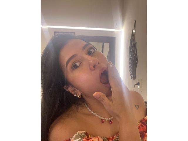 Velka with dark hair, brown eyes, and tan skin, mouth open, tongue out, touching her tongue to her finger, wearing a red and orange dress, pearl necklace, heart tattoo.