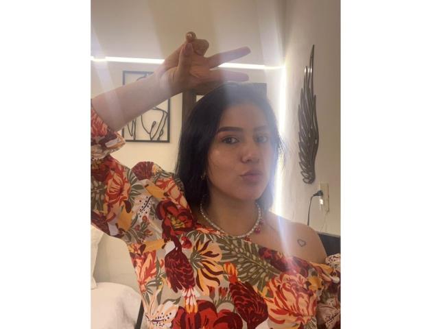 Velka with dark hair, wearing a floral off-shoulder top, making a peace sign with her right hand, has a small heart tattoo on her left shoulder, pouting.