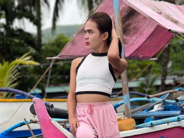 PeachAnderson-hot with dark hair, wearing a white-black sleeveless top and pink shorts, stands by a pink boat, hand on head, looking to the side. Boats and palm trees.