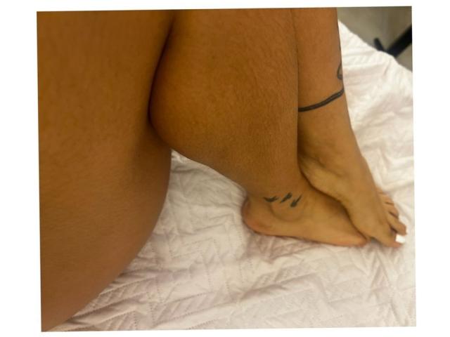 Velka's tan, hairy butt and tattooed ankle with three black lines are on a white, quilted bed. Her feet are crossed at the ankle.