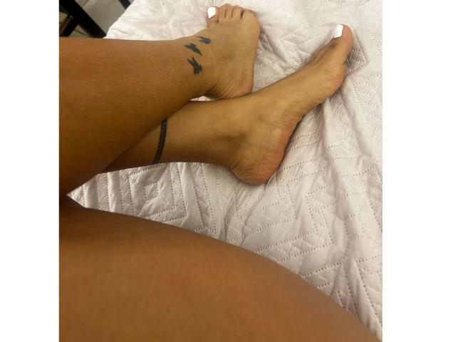 Velka's tanned legs with white-painted toes, a black ankle bracelet, and a small tattoo of three black lines on her left ankle, lying on a white quilted bed.
