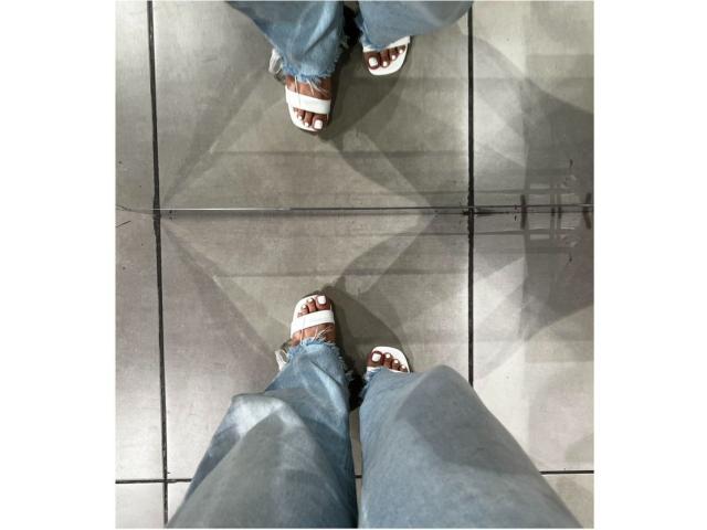 Velka's lower legs and feet, wearing blue jeans and white strappy sandals with brown accents, standing on gray tiled floor, shot from above.