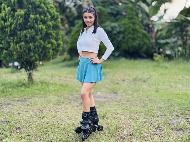 KimmyK with long brown hair, white crop top, blue skirt, and black roller skates, standing on grass, hands on hips, smiling, greenery in background.