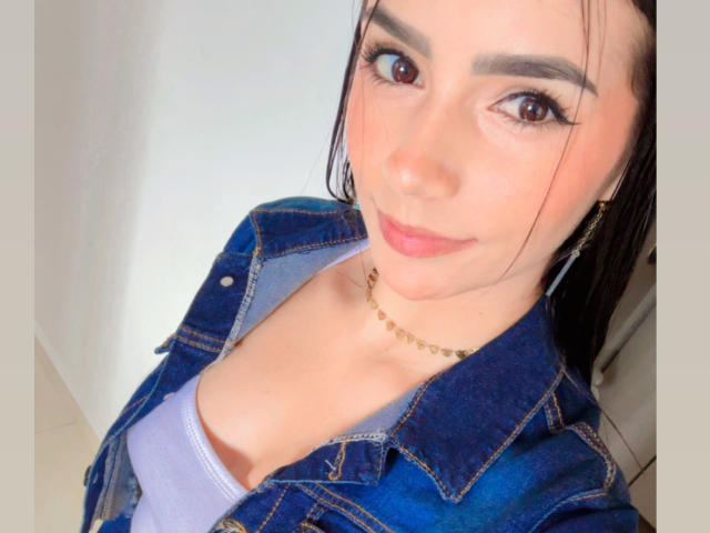 KimmyK with brown eyes, wearing a denim jacket over a purple top, showing her small tits and a gold chain necklace, smiling softly.