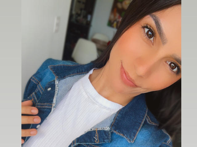 KimmyK with brown eyes, smiling, wearing a denim jacket over a white ribbed shirt, holding her jacket collar, medium skin, long dark hair, and subtle makeup.