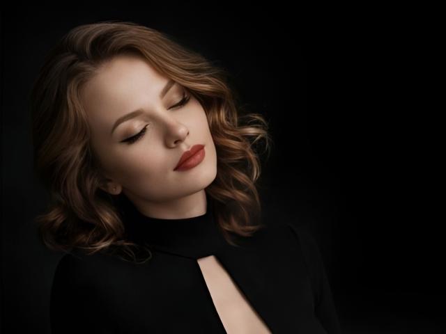 VictoriaAlarie with closed eyes, wavy brown hair, and red lipstick, wearing a black top with a keyhole cutout, has a soft, relaxed expression. Dark background.