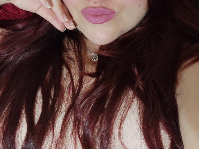 ElizabethFire's face with pink lipstick, dark red wavy hair, and a small necklace. Her hand is touching her cheek, showing off her pink nails. Boobs partially visible.