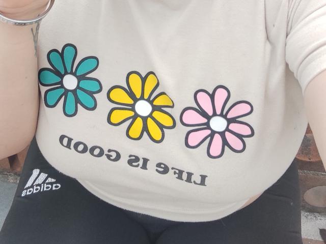 ElizabethFire's tits are covered by a beige shirt with colorful flower designs, yellow, teal, and pink. She's wearing black pants and a silver bracelet.