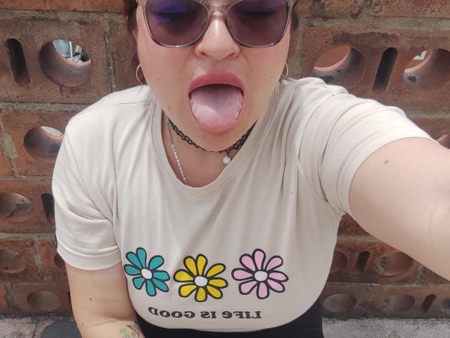 ElizabethFire with tongue out, wearing sunglasses, white t-shirt with colorful flowers, black choker, and hoop earrings, looking up at camera. Her tits are visible under the t-shirt.