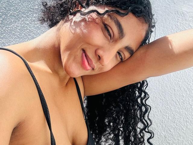 NatalyBlonder has curly black hair, tan skin, and small tits. She's smiling, wearing a black strap top, with her arm raised. Sunlight highlights her face.