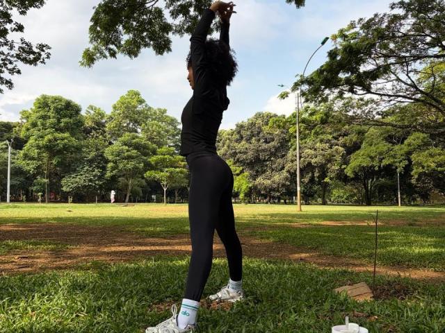 AylaCallowey with curly hair, black long-sleeve top, and black leggings, stretching arms up, standing on grass, wearing white socks and sneakers, in a park.