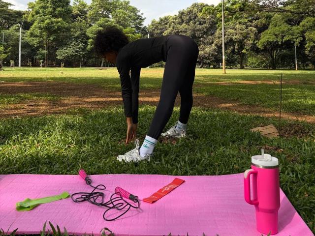 AylaCallowey, black long-sleeve top, black leggings, white sneakers, bending over, pink mat, green and red exercise tools, pink water bottle, grassy park.
