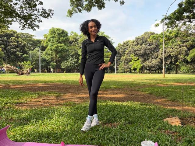 AylaCallowey with curly black hair, wearing a black long-sleeve top and black leggings, standing on grass with one leg raised, white sneakers, in a park.