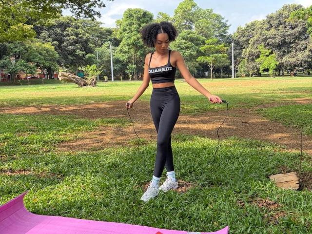 AylaCallowey with curly black hair, wearing a black sports bra and high-waisted black leggings, jumping rope in a grassy park, white sneakers, purple mat in the foreground.