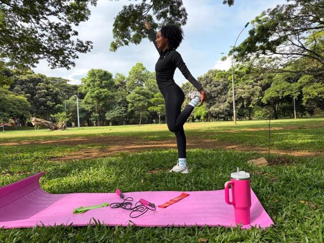 AylaCallowey stands on a pink yoga mat, stretching with one arm raised, wearing black leggings and a long-sleeve top, holding a water bottle, surrounded by green grass.