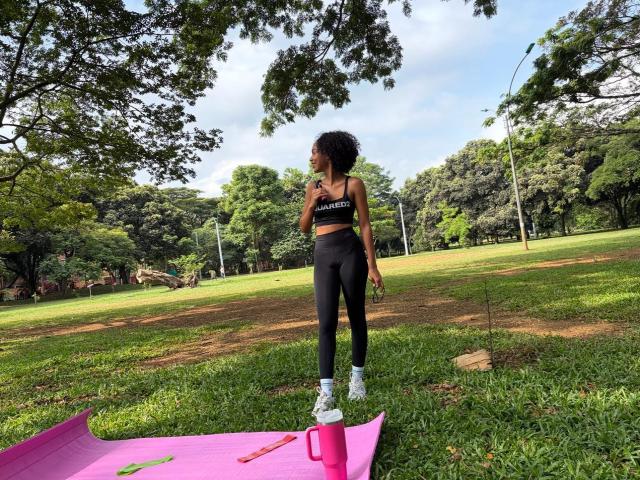 AylaCallowey stands on a pink yoga mat in a grassy park, wearing black sports bra and high-waisted leggings, white sneakers, with a pink water bottle on the mat.