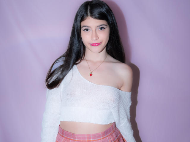 RachelHottie with long black hair, white off-shoulder sweater, pink plaid skirt, red lipstick, and a red necklace, smiling at the camera. Her tits are partially visible.