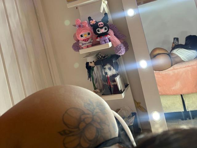 AlessiaFerry's tattooed butt in the foreground, reflection in mirror showing her ass in black thong, stuffed toys on shelf, dress hanging, and lit mirror in bedroom.