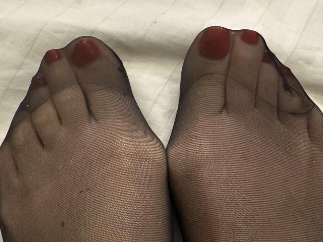 NancyChaude's feet in sheer black stockings, toes slightly spread, with red-painted toenails. The texture of the stockings is visible, showing fine lines and slight dark marks. The feet are centered, toes pointing upwards, with a close-up view emphasizing the toes and nails. The background is a white, slightly textured surface. The image has a realistic, photographic style with natural lighting.