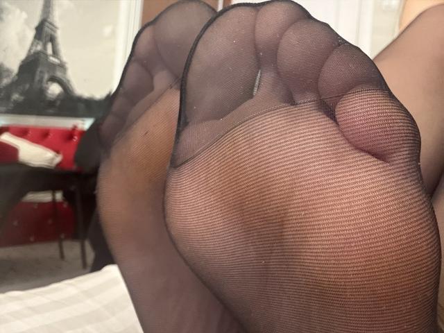Close-up of NancyChaude's feet in black sheer stockings. Her toes are slightly curled, showing the textured fabric of the stockings. The focus is on the soles, with the toe tips and heel visible. The background includes a blurred image of the Eiffel Tower. The composition emphasizes the texture of the stockings and the natural curves of her feet. The lighting is even, highlighting the subtle shadows and contours. The image uses a shallow depth of field to keep the focus on the feet.