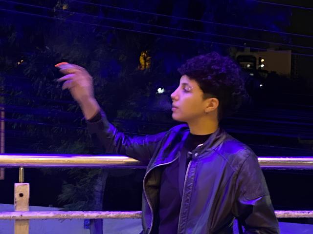 AlinaRicker with curly black hair, wearing a black shirt and shiny leather jacket, raises a hand, illuminated by blue and purple lights, in front of a metal railing.
