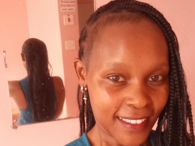 IvyTheeGodess has dark skin, long braided hair, and a small mole under her right eye. She's smiling with red lipstick, wearing a blue top, and earrings.