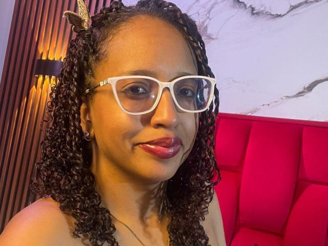 ArtemisaS has curly black hair with red accents, white glasses, and shiny pink lips. She's wearing a red headband and smiling slightly. Her brown skin looks smooth.
