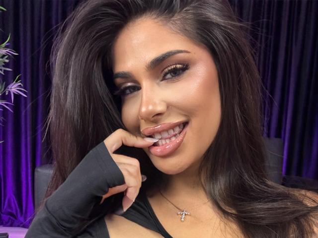 BabeBlueSin with long dark hair, glossy pink lips, and a finger in her mouth, wearing a black top and a cross necklace, smiling seductively.