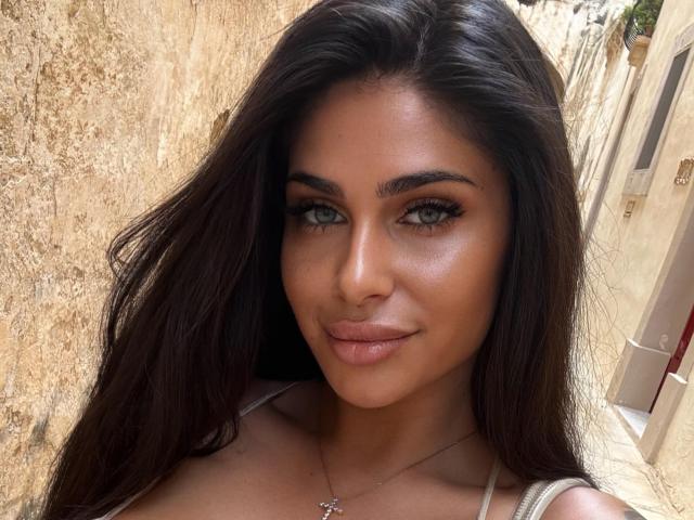 BabeBlueSin has long dark brown hair, green eyes, and full pink lips. She's wearing a white top and a silver necklace, smiling softly. Her tits are slightly visible.
