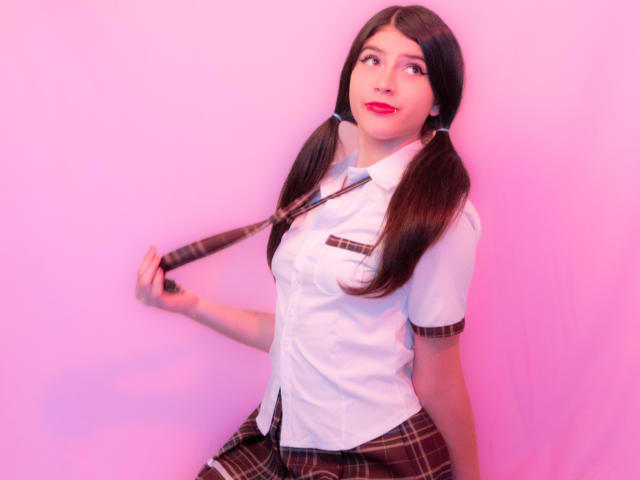 RachelHottie with pigtails, white shirt, brown plaid skirt, and matching tie, holding her tie against a pink wall, smiling with red lipstick, showing her tits slightly.