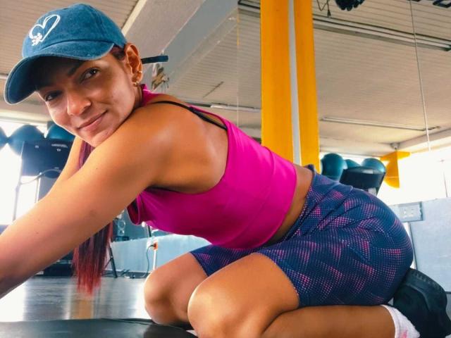 HannaPretty with tan skin, red ponytail, and blue cap, wearing a pink sports bra and blue mesh shorts, kneeling on a gym mat, showing her ass and a smile.