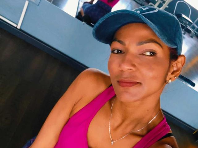 HannaPretty with tan skin, brown eyes, and small tits, wearing a pink top, blue cap, and silver necklace, smiling slightly. She's indoors, with a wooden wall behind.