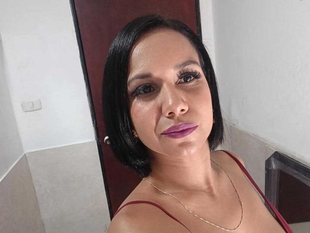 EvelinFoster has shiny skin, black bob, purple lipstick, long eyelashes, gold necklace, and red top, smiling at the camera with visible tits and a cute ass.