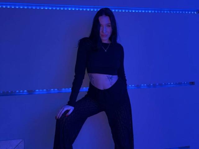 NikkiGoldX with long black hair, wearing a black crop top and black patterned pants, showing a tattoo on her right side, standing with one leg raised, blue-lit.
