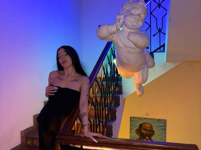 NikkiGoldX with long black hair, bare boobs, tattoos, black strapless dress, standing by staircase, holding her ass, with a white baby statue and colorful stairs in background.