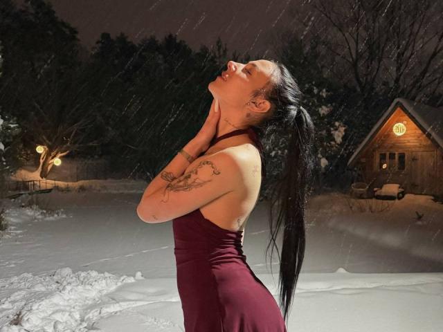 NikkiGoldX stands in a snowy night, rain falling. She's in a maroon dress, backless, with long black hair in a ponytail. She tilts her head back, eyes closed, hands on her neck. Her tattoos are visible on her left arm. A wooden house with a lit window is in the background. The snow and rain create a soft glow on her skin. The image has a realistic, slightly dramatic style.