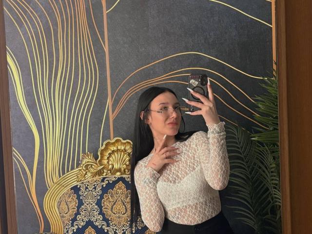 NikkiGoldX with long black hair, glasses, white lace top, holding phone, blue patterned chair, gold headrest, green plant, taking mirror selfie, showing off manicured nails.