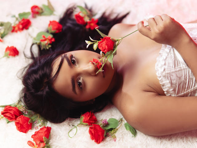 MariamLara lies on a white fluffy rug, holding a red rose to her mouth, wearing a white lace strapless dress, with more red roses scattered around her.