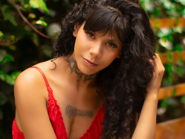 StormyWest with curly black hair, red lace top, tan skin, and a tattooed chest, touching her hair, showing her tits, brown eyes, and a slight smile.