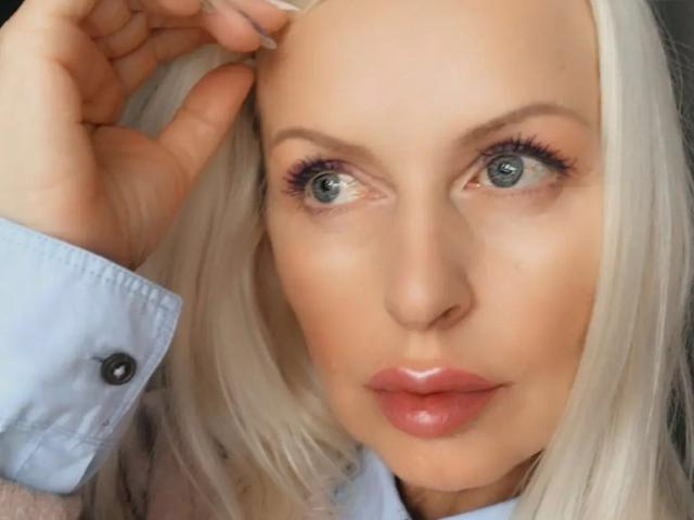 RosalineRena has platinum blonde hair, blue eyes, and glossy pink lips. She's wearing a light blue shirt with black buttons, touching her hair with her right hand.