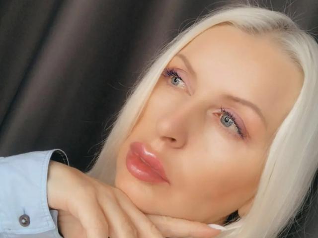 RosalineRena's face with blonde hair, pink glossy lips, blue eyes, and light skin, wearing a blue shirt, her hand gently touching her chin.