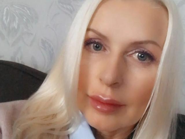 RosalineRena has blonde hair, blue eyes, and glossy pink lips. She's wearing a light pink jacket over a white shirt. Her face is slightly angled to the camera.