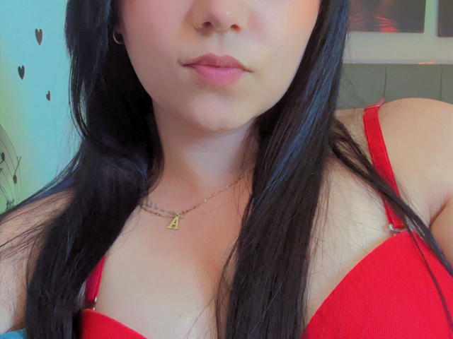 Kkatalina with dark hair, red bra, and gold necklace, showing her boobs and pouty lips, wearing small earrings. Her skin is fair, and she's looking at the camera.